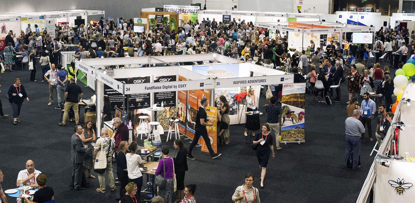 Large indoor event with crowds networking and visiting exhibition booths in a convention center.