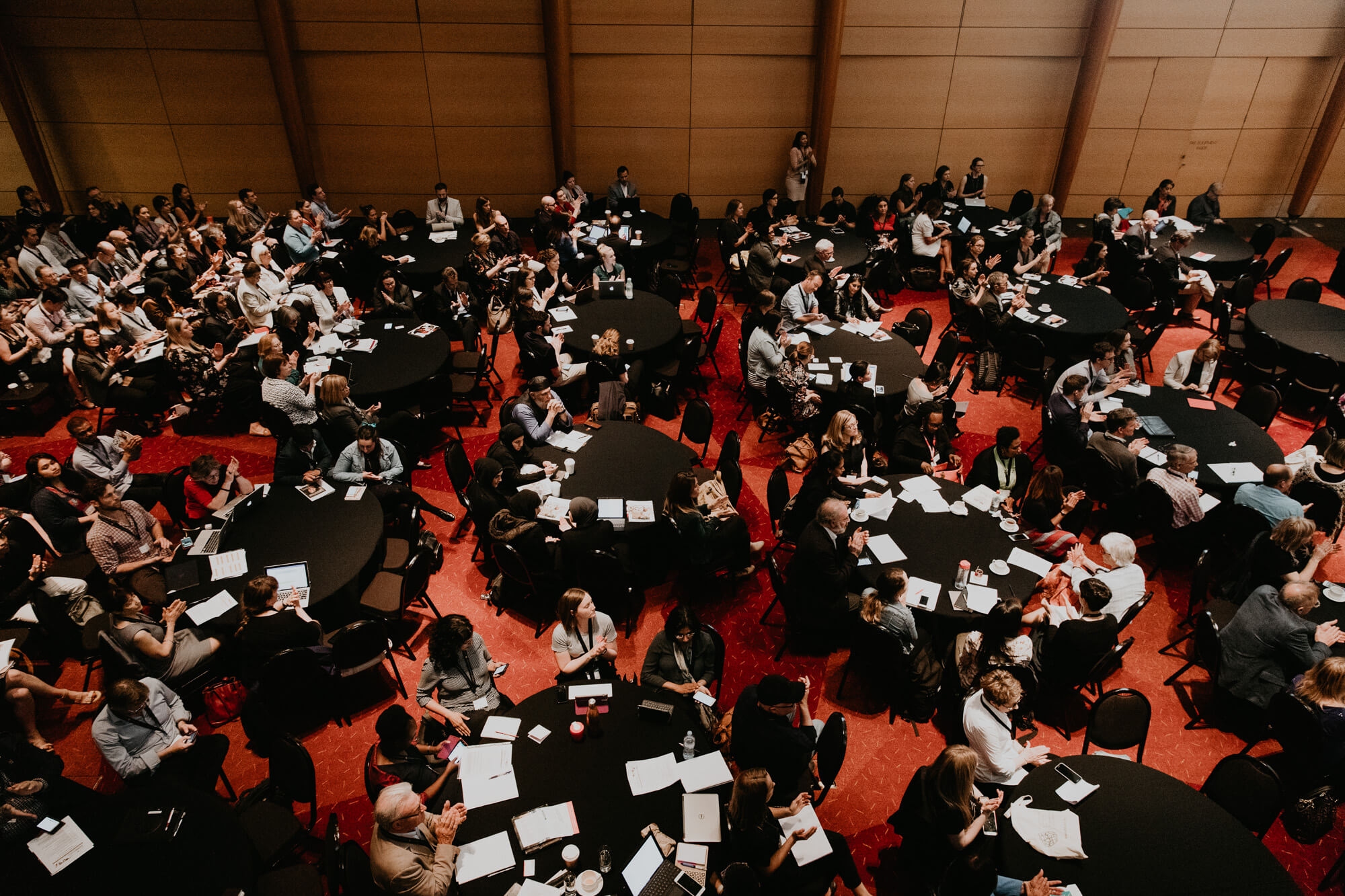 Overhead view of a large group of people seated at round tables during a conference or seminar in a spacious room.