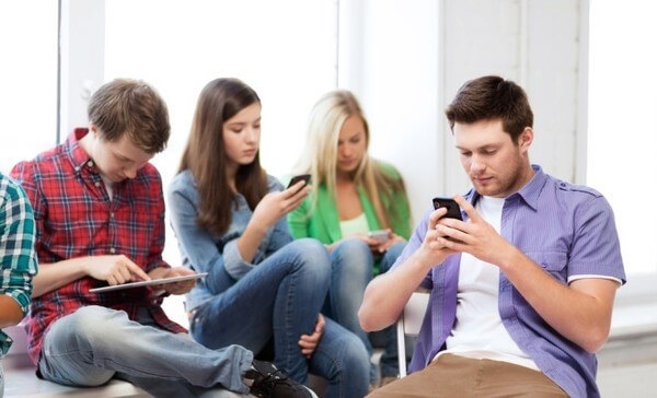 Four young adults sitting together, focused on their smartphones and a tablet in a bright indoor setting.