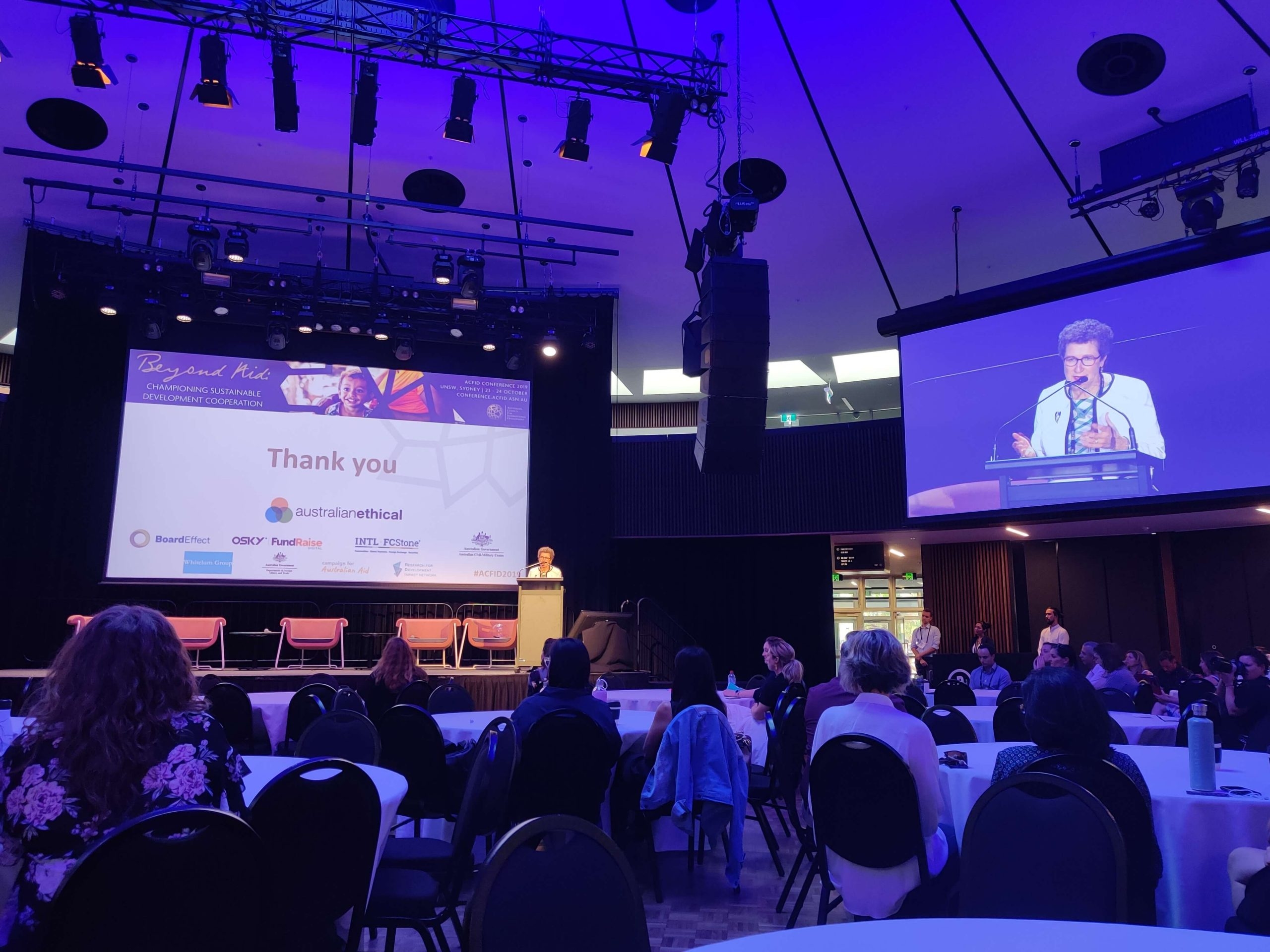 Audience seated at a conference listening to a speaker at a podium with a "Thank you" slide projected on stage.