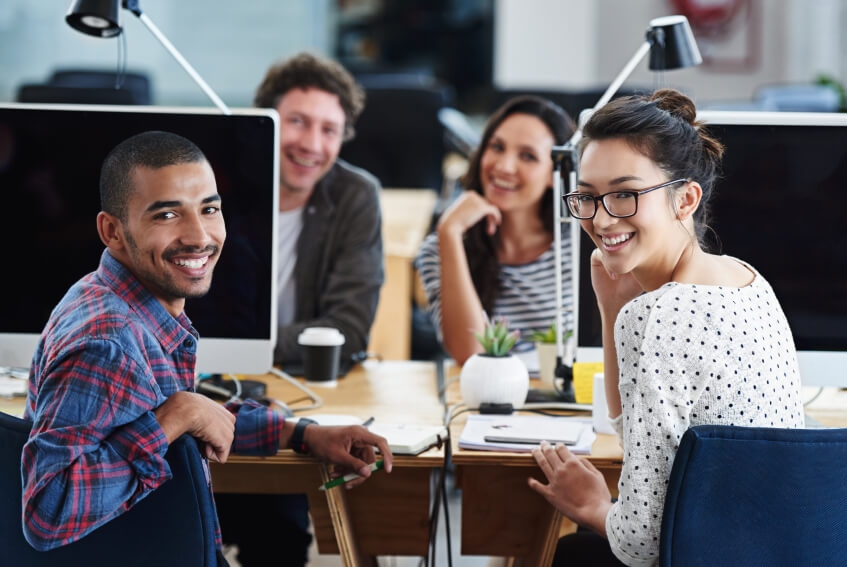 Four diverse young adults smiling and sitting around a desk with computers in a modern office setting.