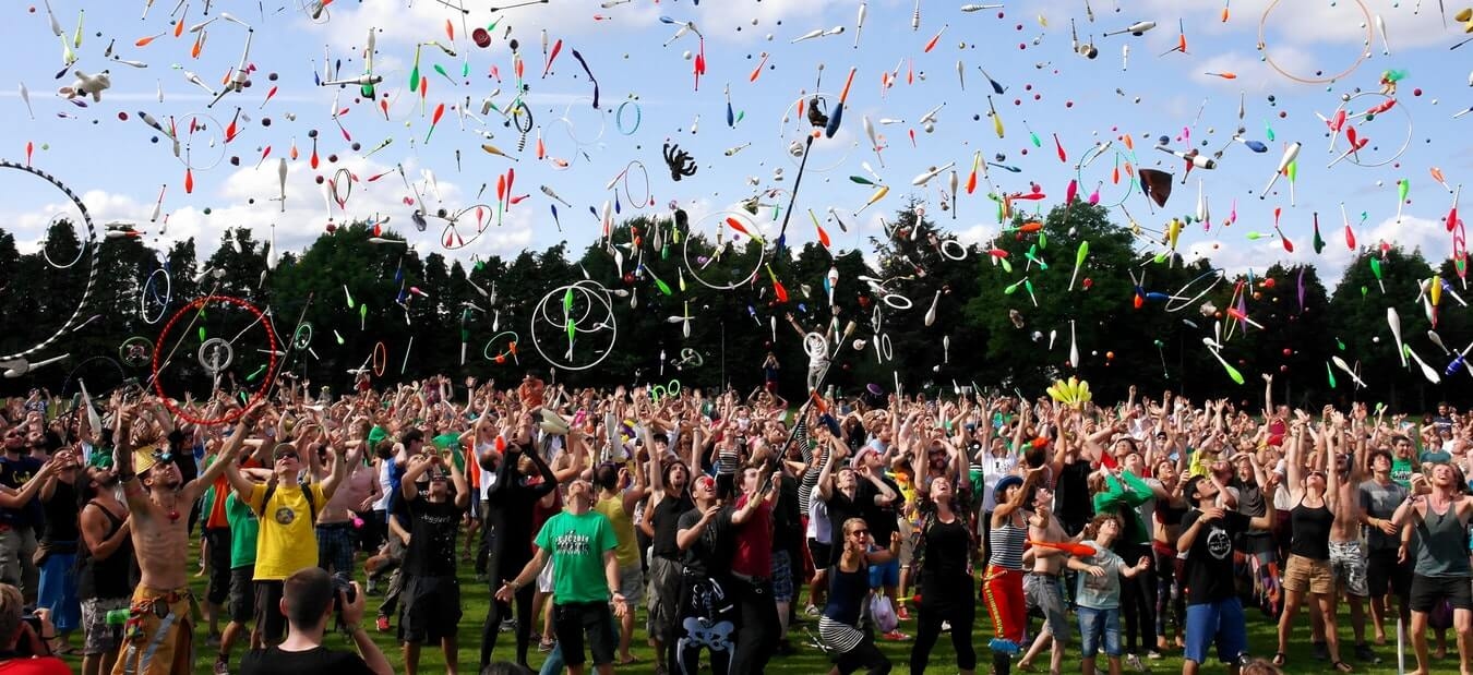 A large crowd outdoors throwing juggling clubs and rings into the air against a blue sky and tree backdrop.