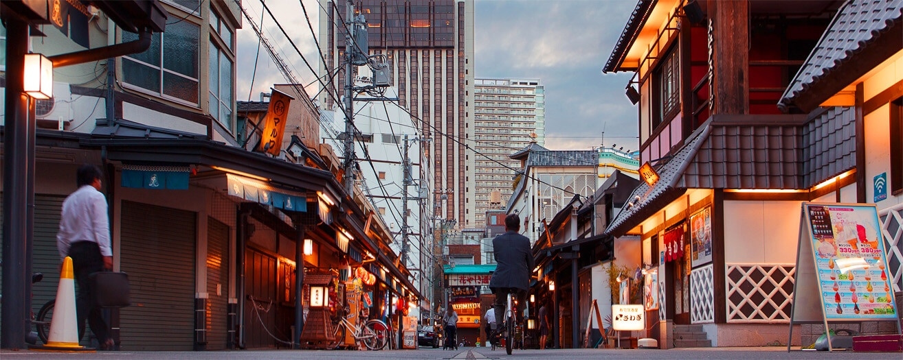 Evening street scene in a Japanese city with traditional buildings, warm lights, and people walking and cycling.