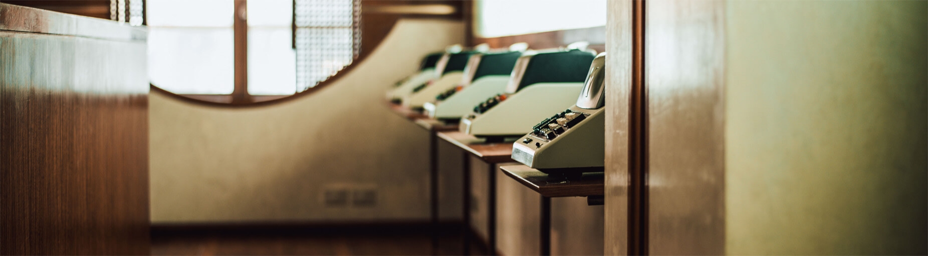 A row of vintage call center telephones on wooden desks in a softly lit room with patterned windows.