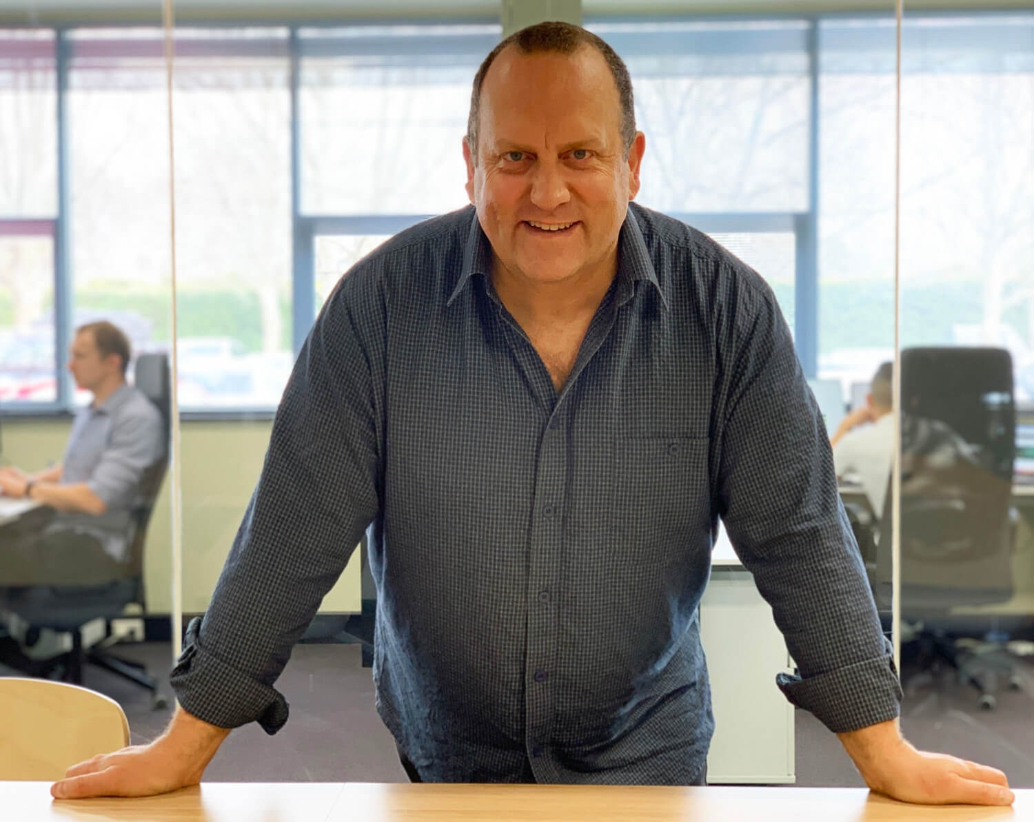 Man smiling and leaning on a wooden table in a modern office with people working in the background.