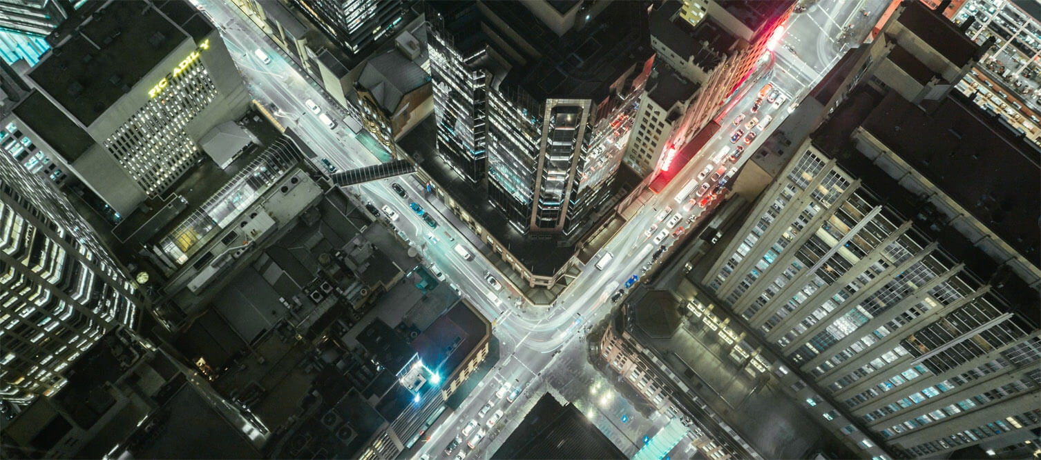 Aerial view of a busy city intersection at night with illuminated buildings and streaks of moving traffic lights.