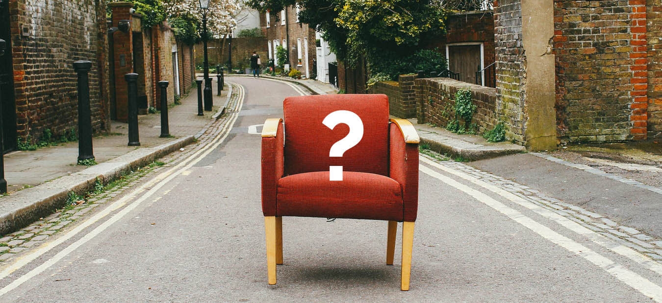 Red armchair with a white question mark placed in the middle of an empty narrow street between brick buildings.