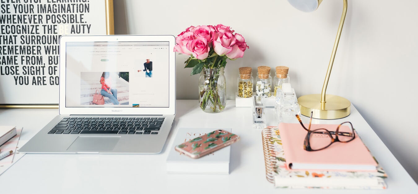 White workspace with a laptop, pink roses in a jar, stationery, a gold lamp, and glasses resting on notebooks.