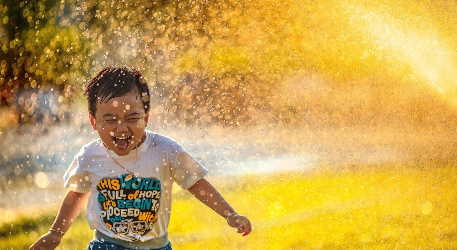 A joyful child wearing a white t-shirt plays in water droplets under golden sunlight.