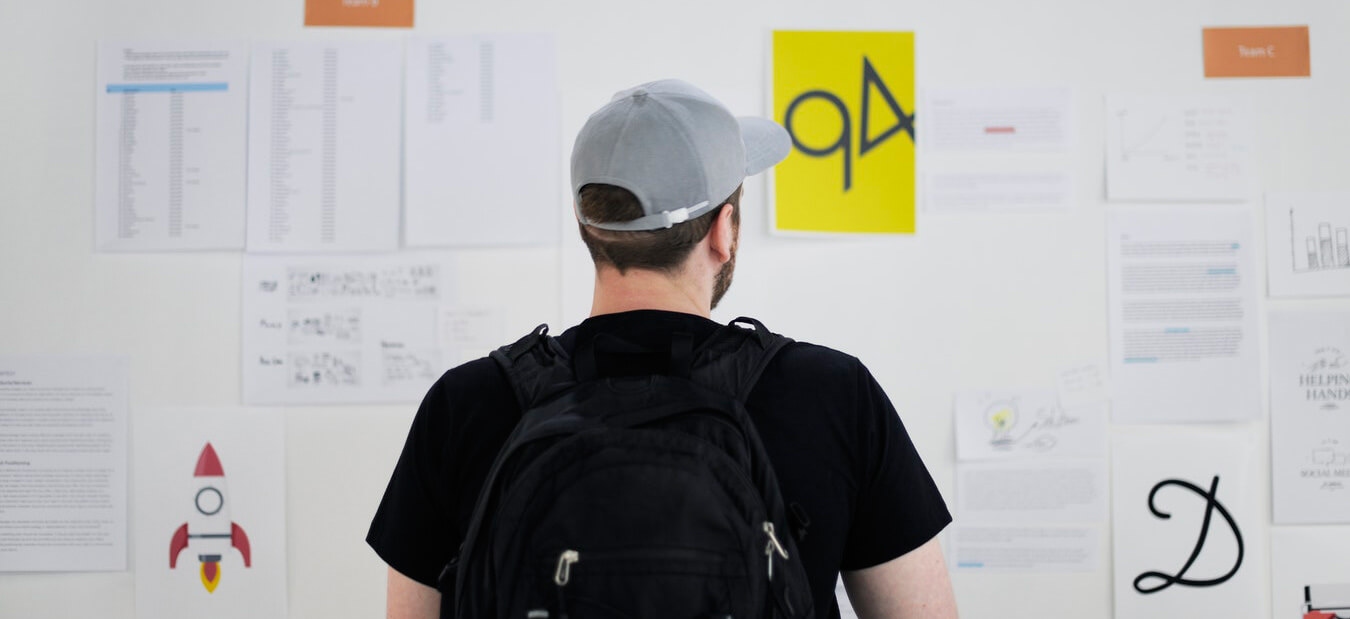 Man wearing a gray cap and black backpack looks at a wall covered with various papers and colorful posters.