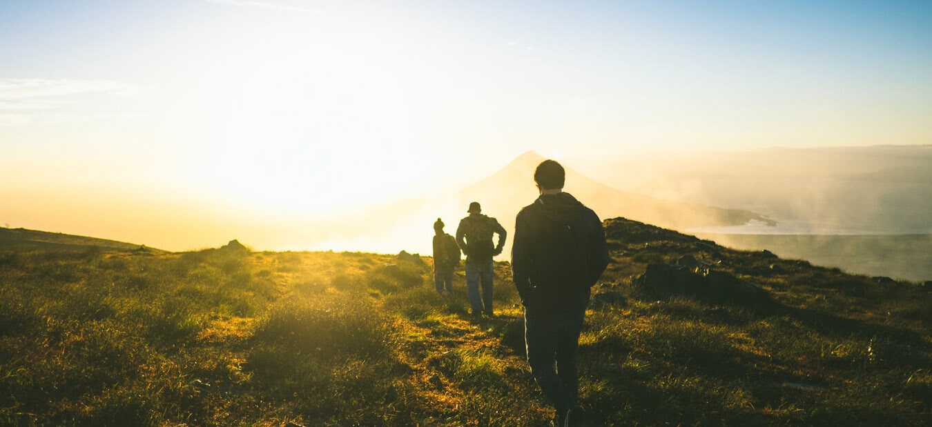 Three hikers walk toward a bright sunrise over a mountainous landscape with grassy terrain and mist.