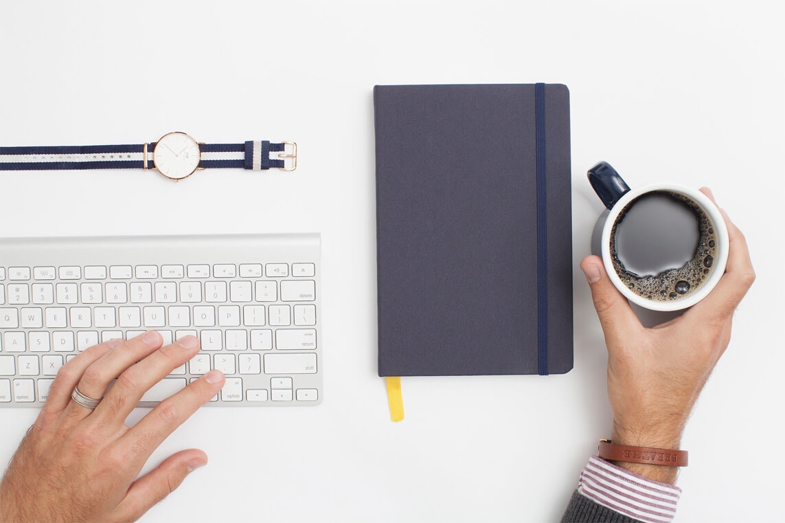 Hands with a keyboard and coffee cup, with a watch and notebook on a white desk.