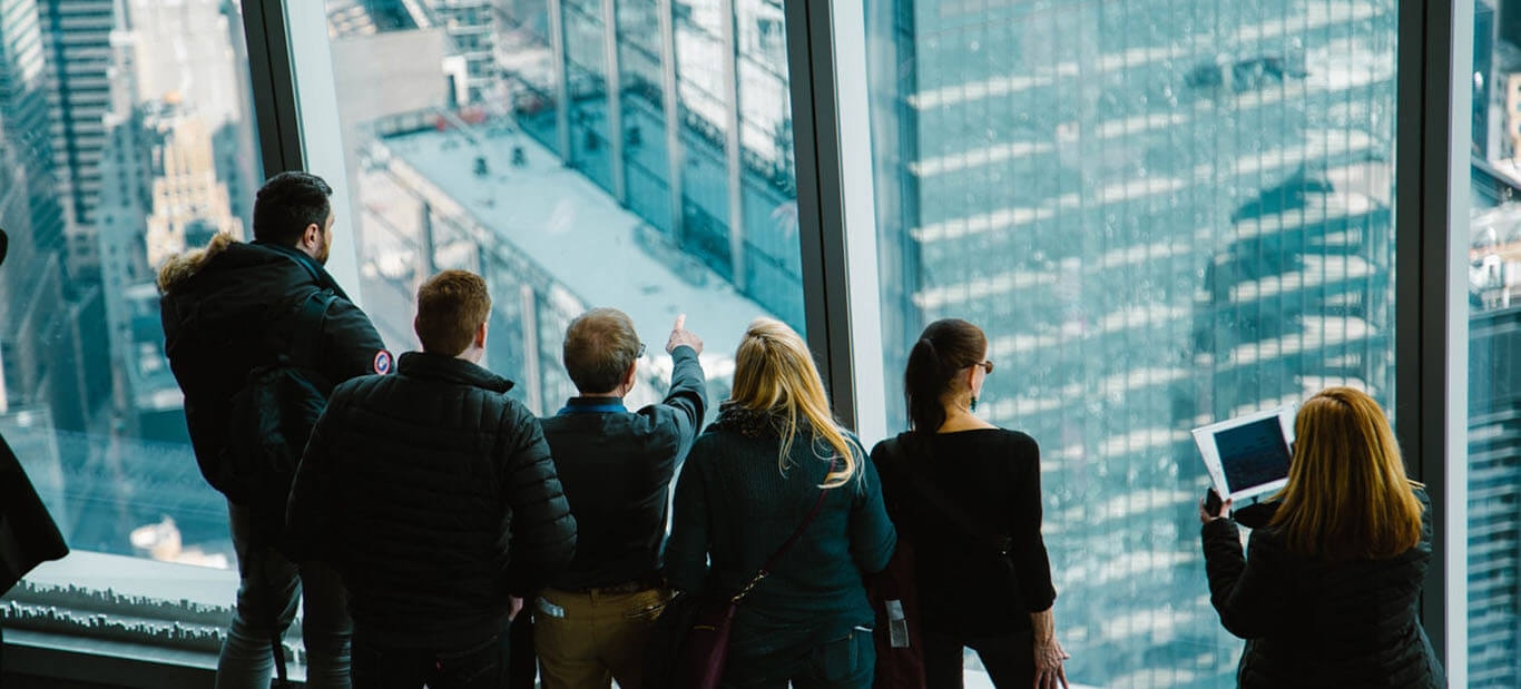 Group of people wearing jackets observing cityscape from a high-rise building with large glass windows.