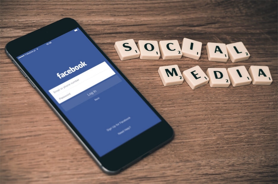 Smartphone displaying Facebook login screen beside Scrabble tiles spelling "SOCIAL MEDIA" on wood.