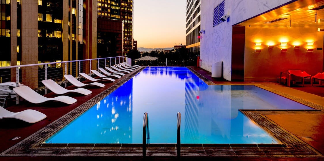 Rooftop swimming pool at dusk with modern lounge chairs, city skyline reflections, and warm wall lighting.