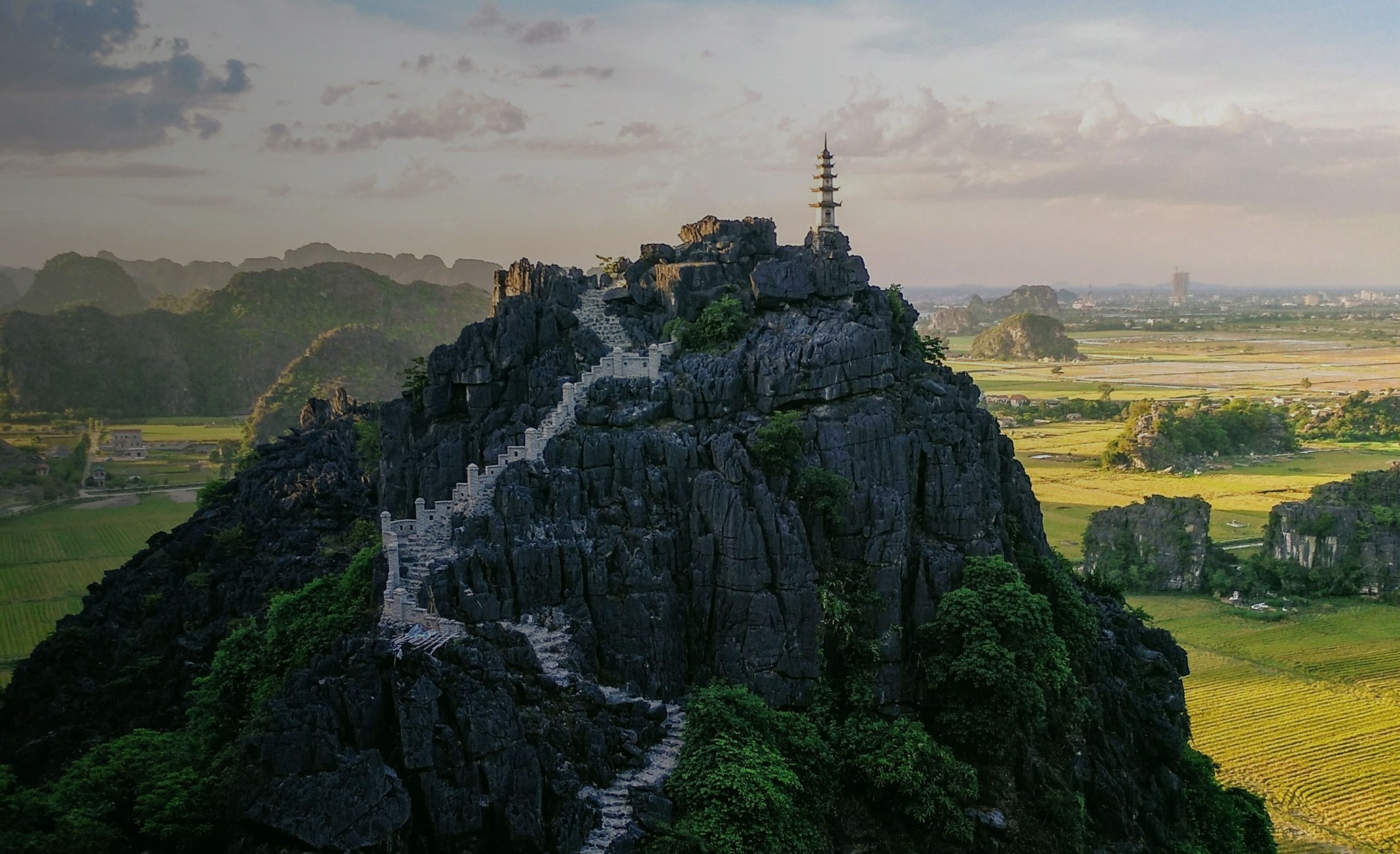 Stone staircase winding up a rocky hill with a pagoda on top, overlooking green fields and distant mountains at sunset.