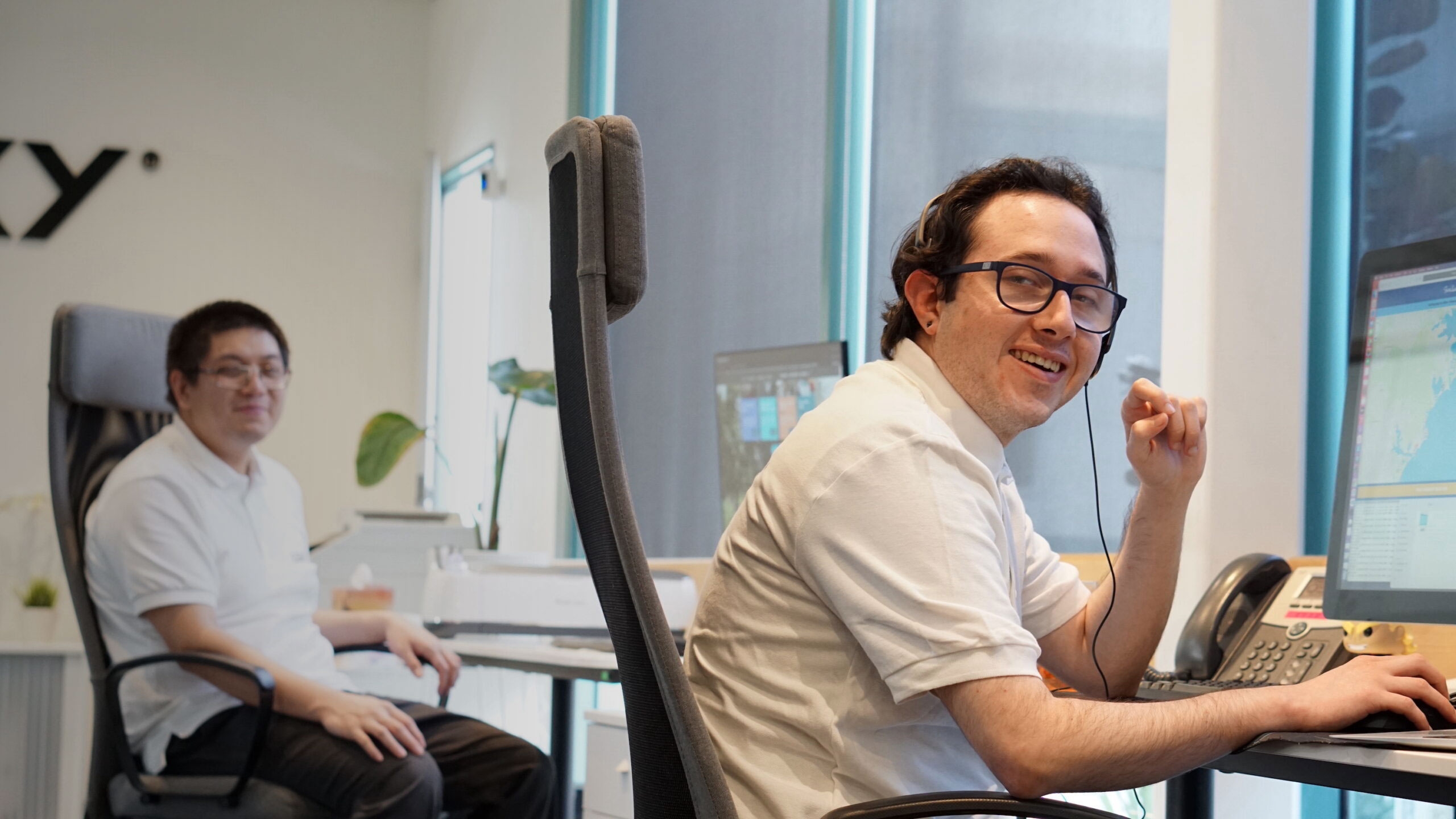 Two men in white shirts working at desks with computers in a bright office space with large windows.
