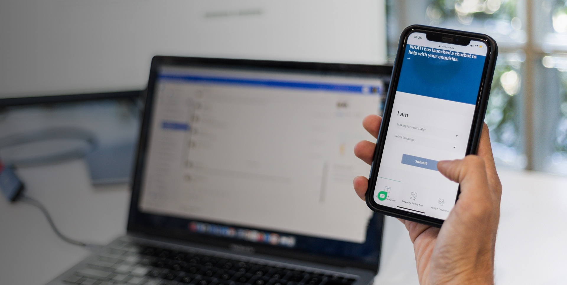 Hand holding smartphone displaying NAATI chatbot screen in front of a blurred laptop on a desk.