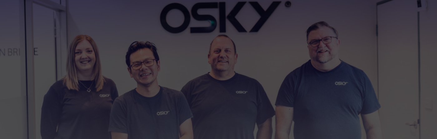 Four smiling coworkers wearing matching black OSKY t-shirts standing side by side against a white office wall with the OSKY logo.