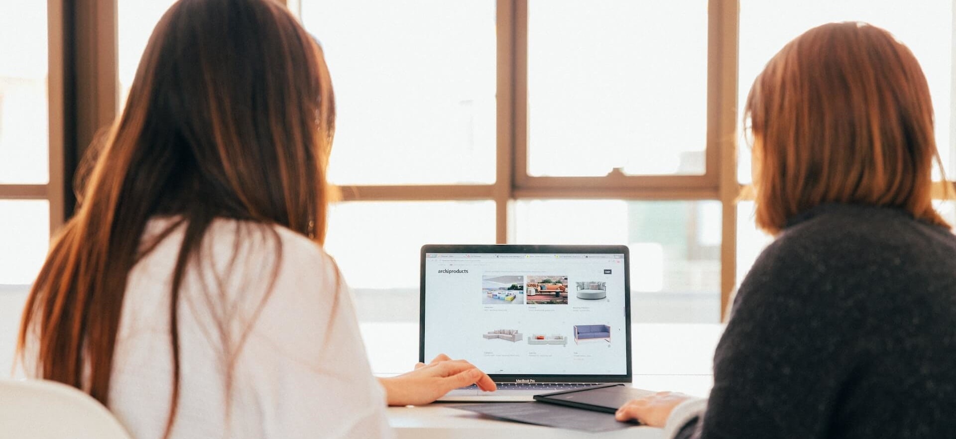Two women viewing furniture options on a laptop at a bright, modern workspace.