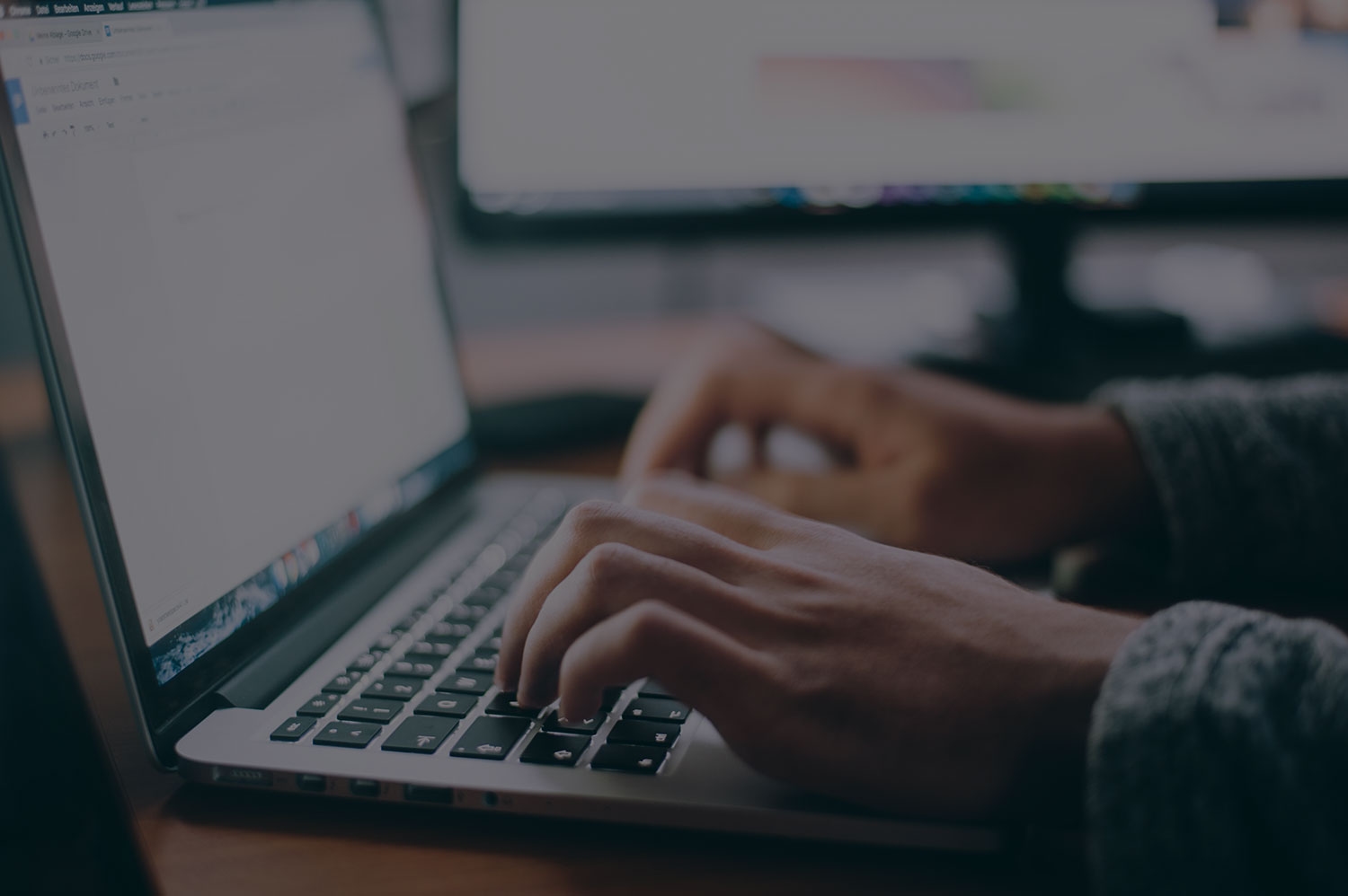 Hands typing on a laptop keyboard with a document open on the screen in a dimly lit workspace.