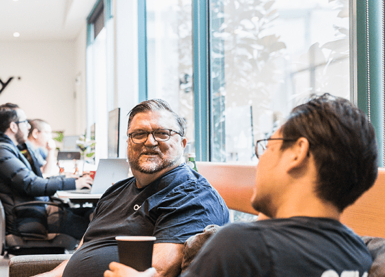 Two men wearing glasses chatting in a bright office lounge with laptops and people working in the background.