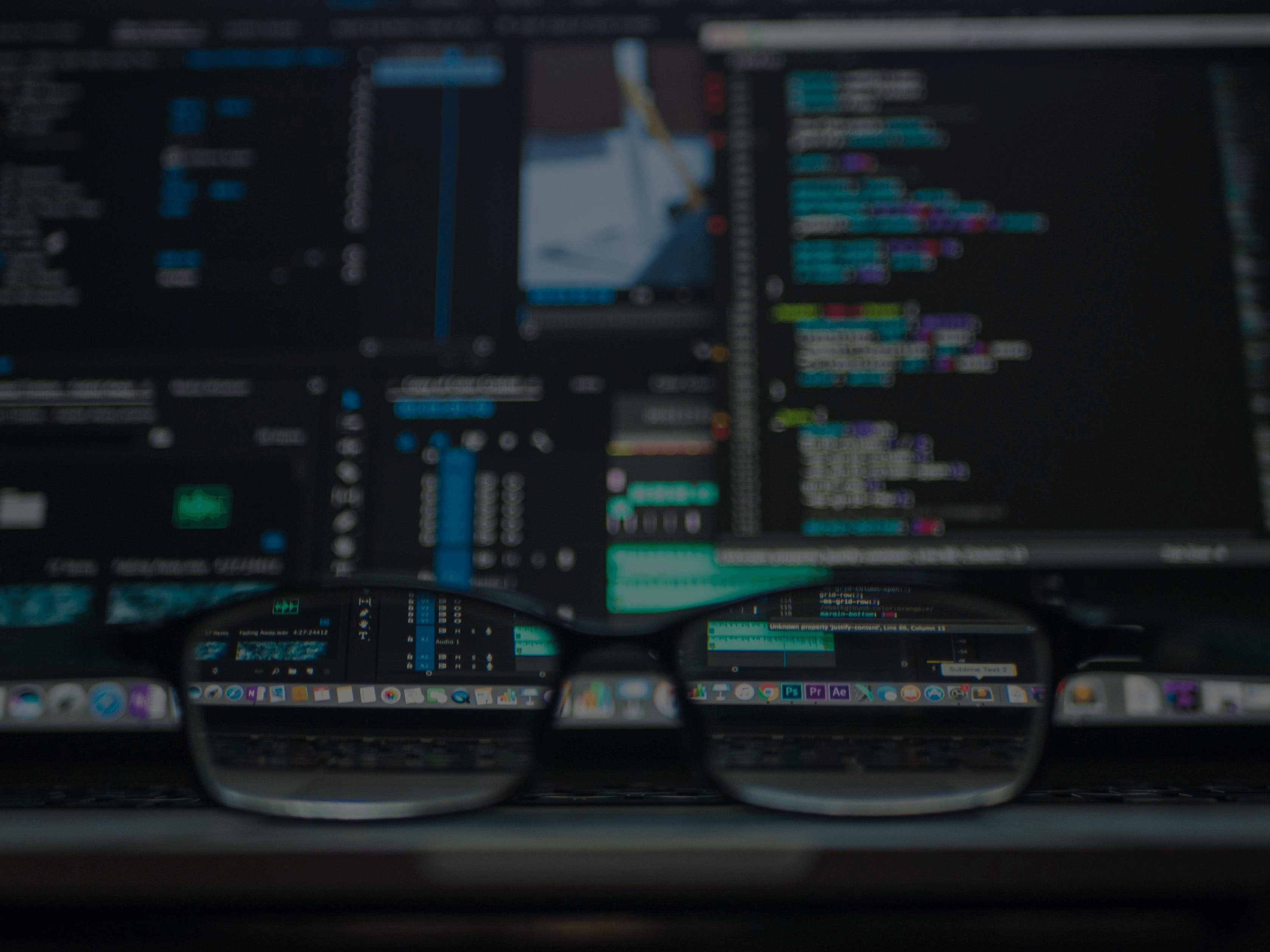 Glasses on laptop keyboard, with computer screen showing blurred and clear coding and software interfaces.