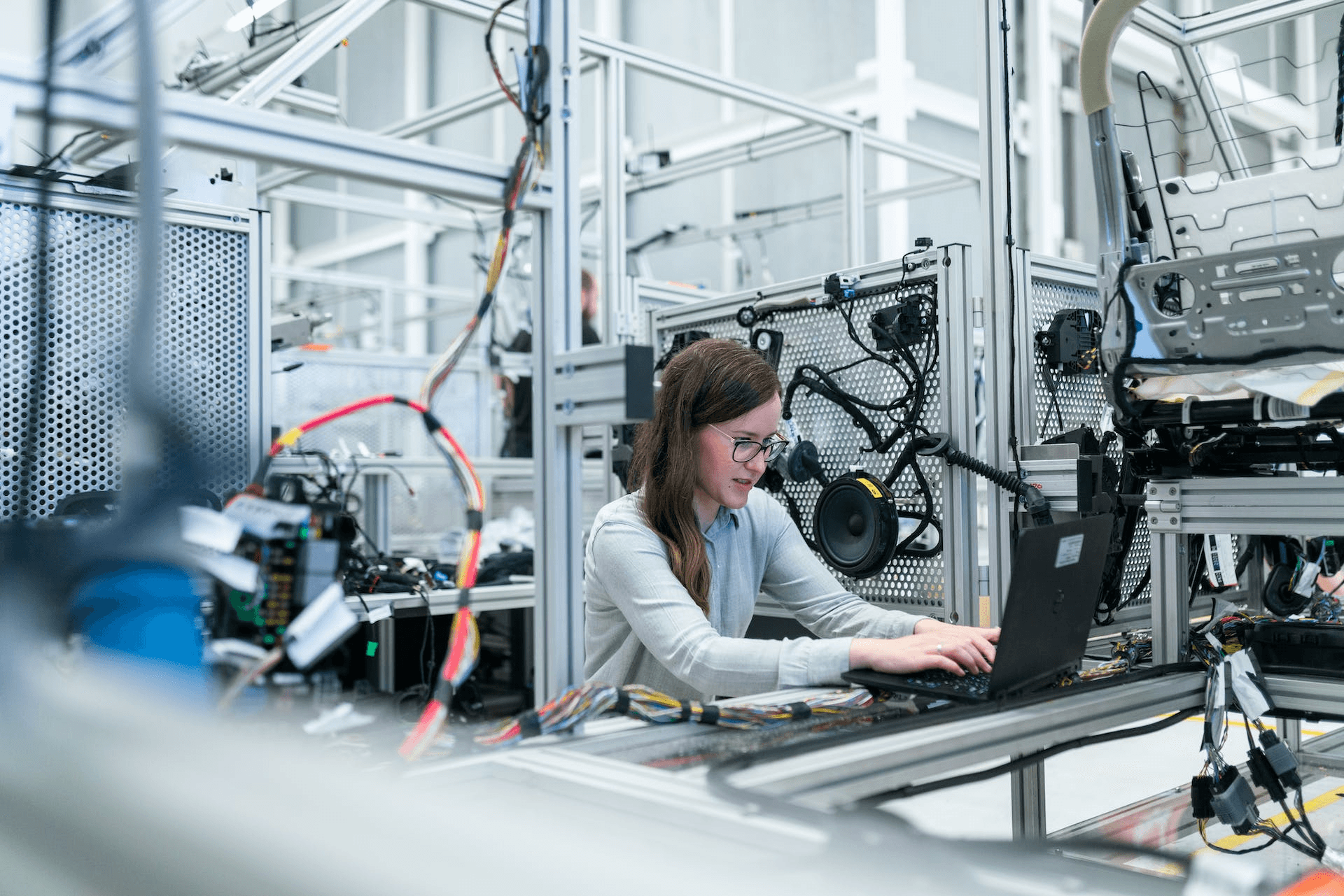 Woman using a laptop inside a vehicle manufacturing setup with wiring and car seat assembly.