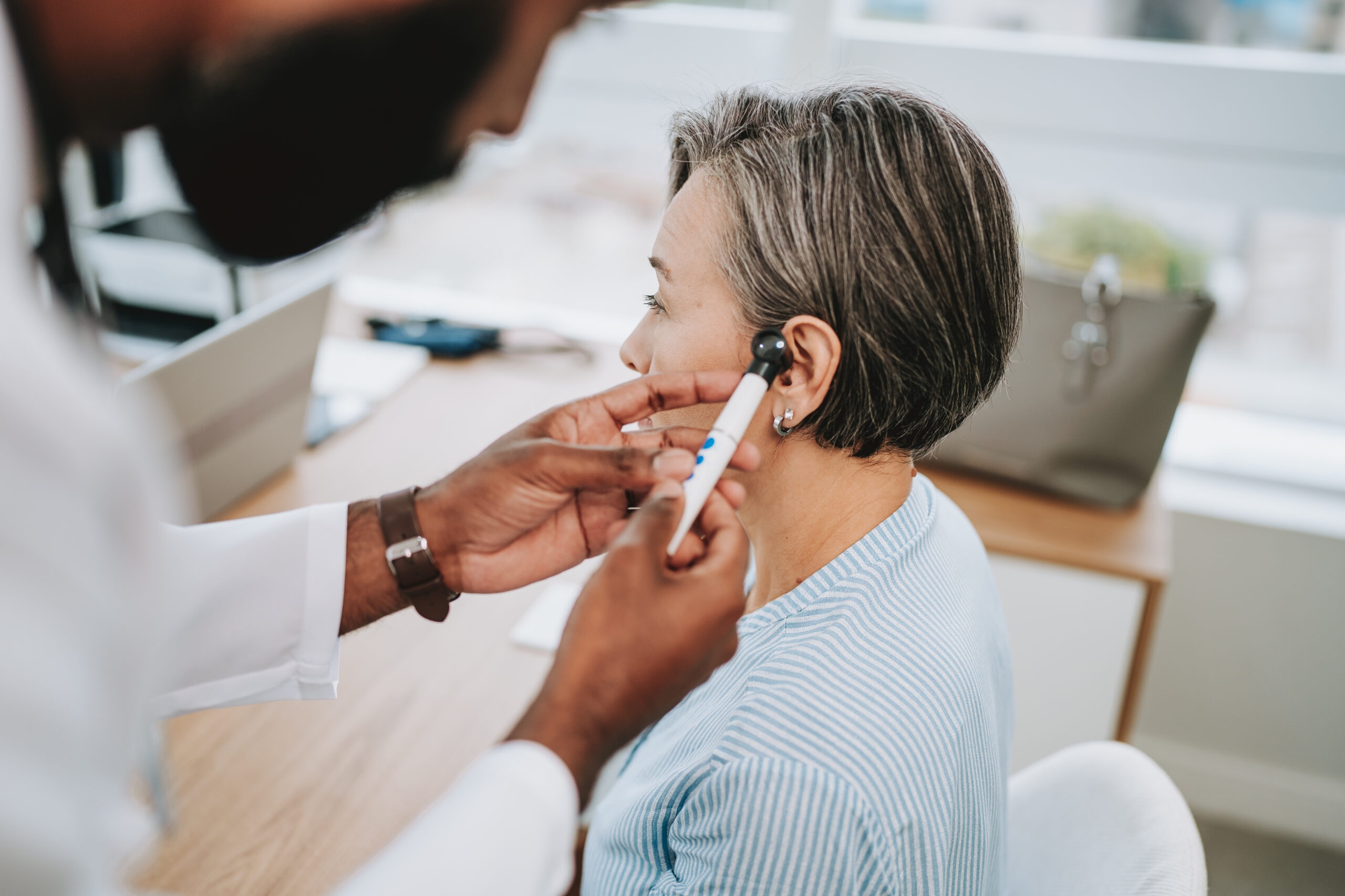 Doctor examining woman's ear with otoscope in a bright medical office.