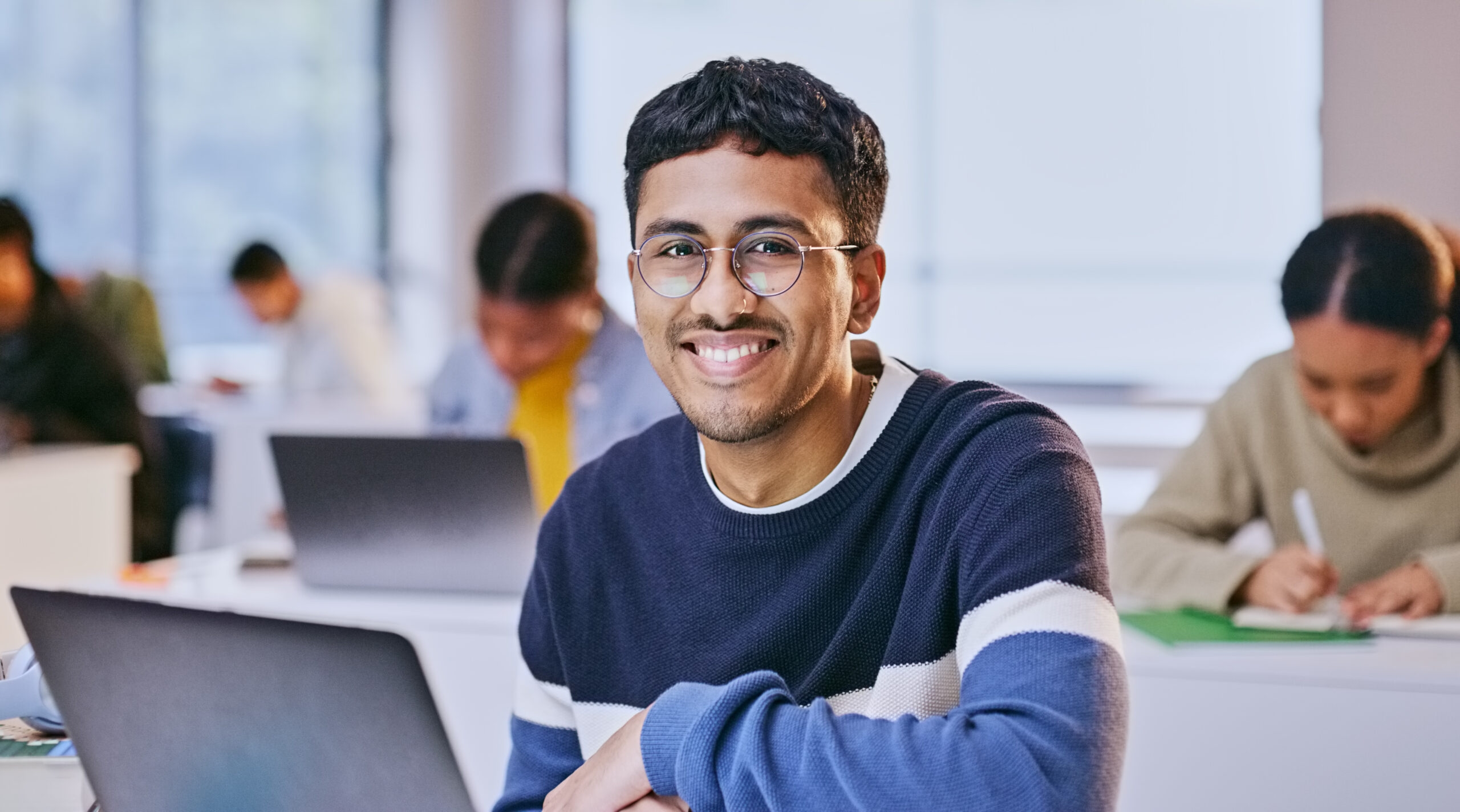 Young man wearing glasses and a striped sweater smiling with a laptop in a classroom.