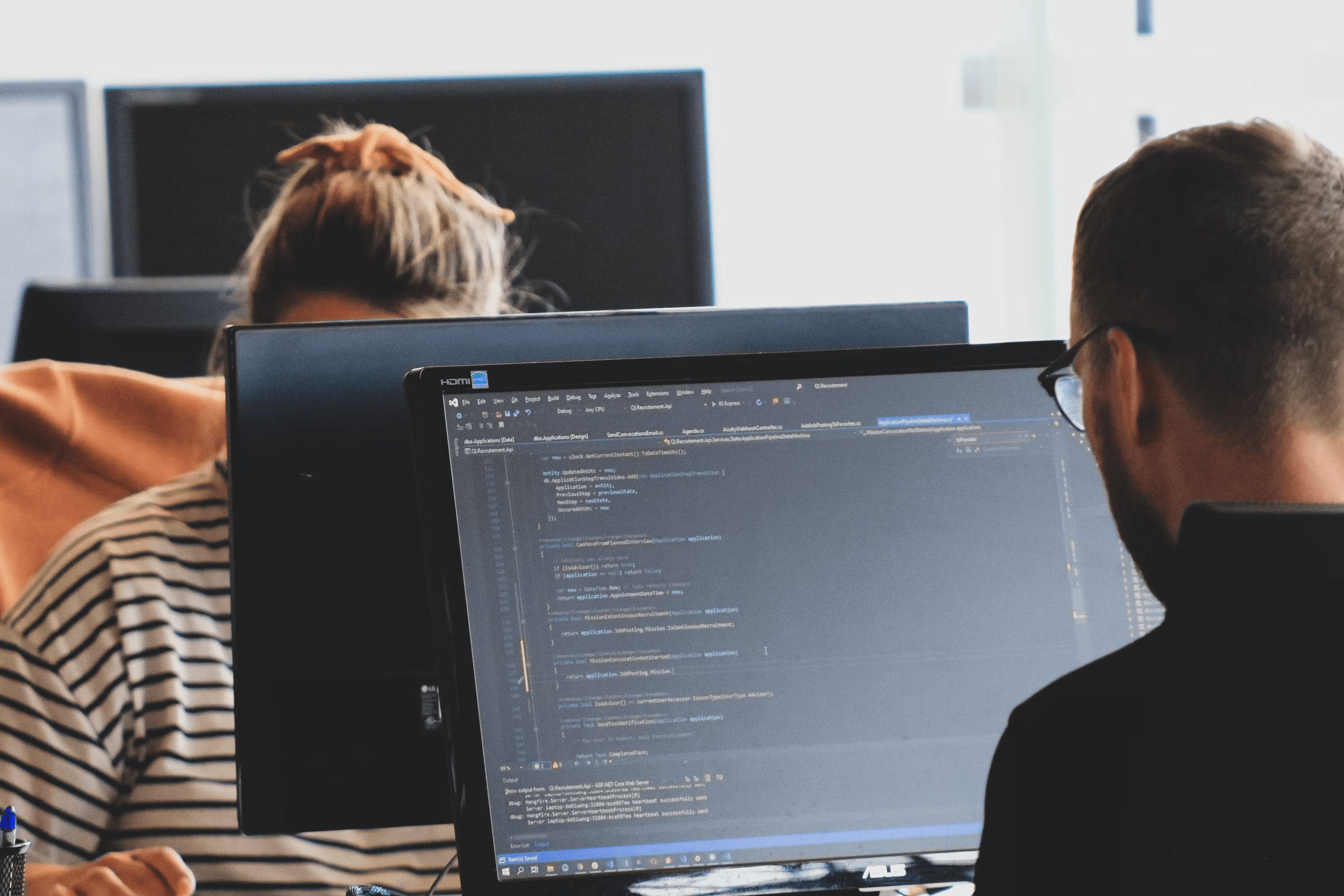 Two people working at computers, one with code displayed on screen, in an office setting.
