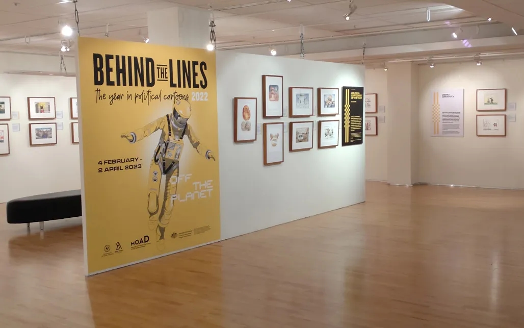 Gallery interior showing "Behind the Lines" political cartoon exhibit poster and framed artworks on white walls.