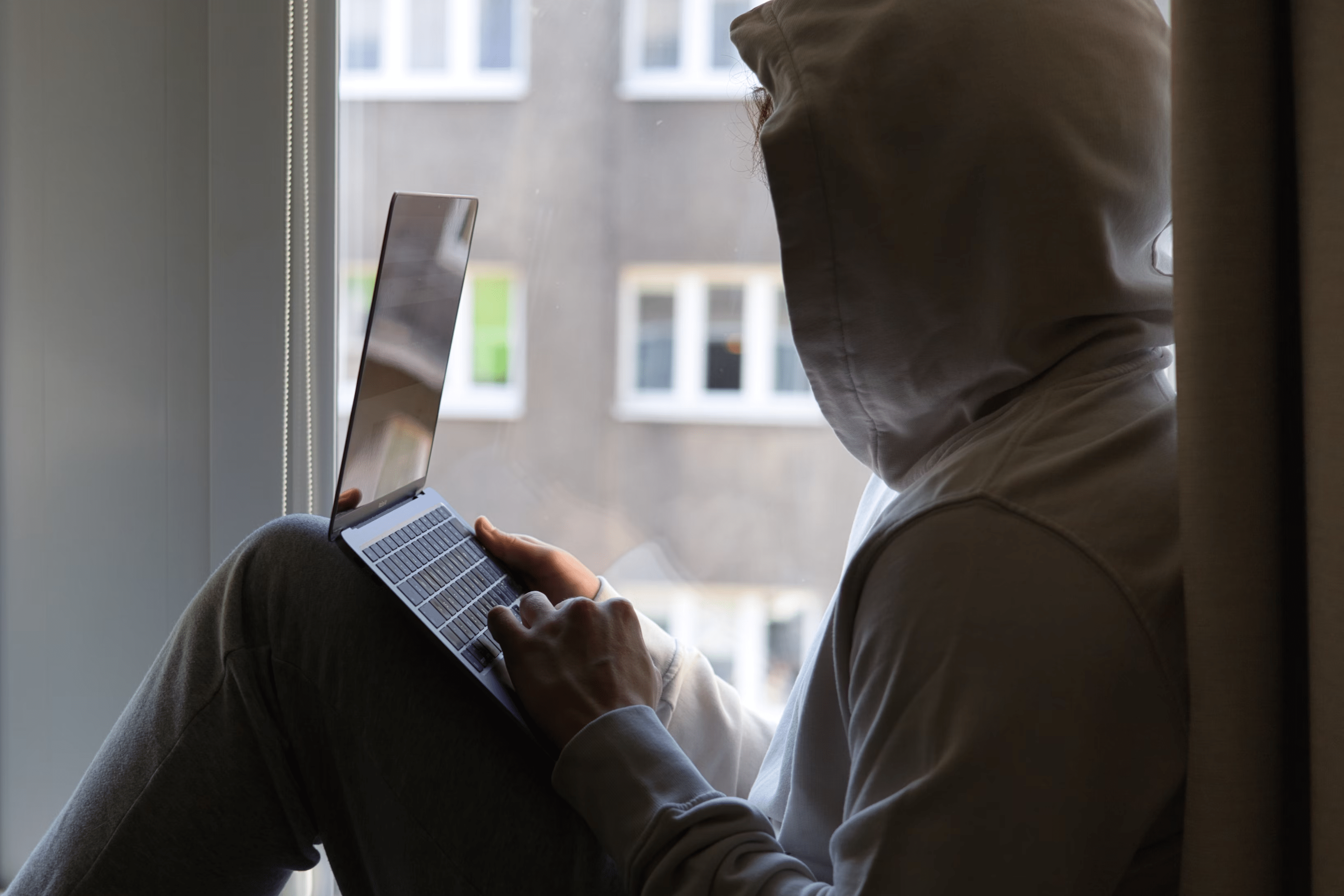 Person in hoodie using laptop near a window with an outside building visible in background.