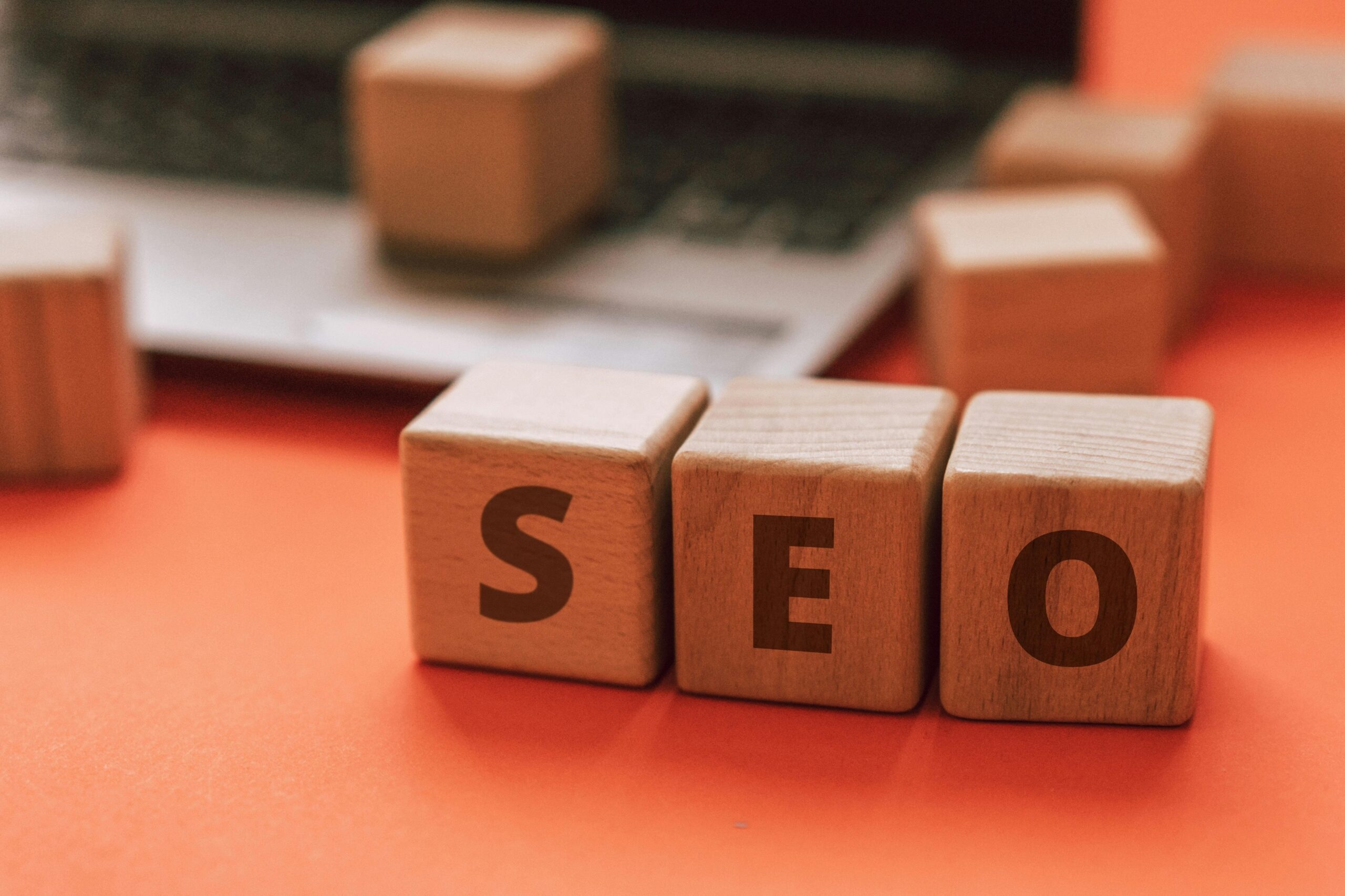 Wooden blocks spelling "SEO" on an orange surface with a blurred laptop in the background.