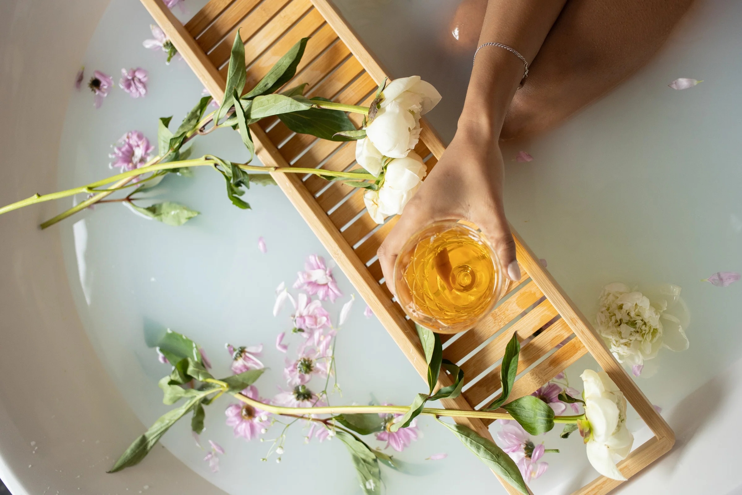 Hand holding a glass of amber liquid over a bath tray with white and pink flowers in milky water.