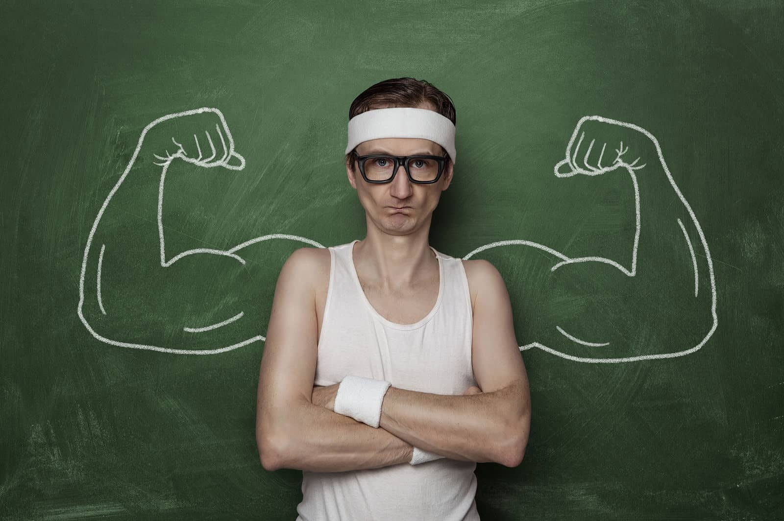 Man wearing glasses and a headband stands with crossed arms in front of a chalkboard with drawn muscular arms flexing.