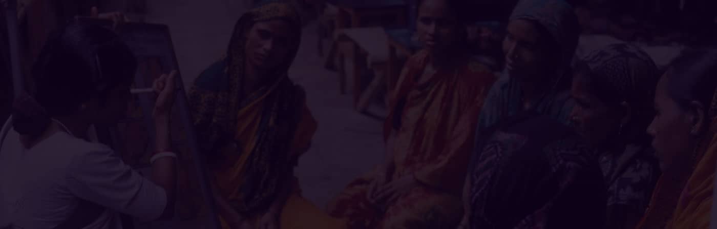 A group of women attentively listening to another woman teaching using a visual aid in an outdoor setting.