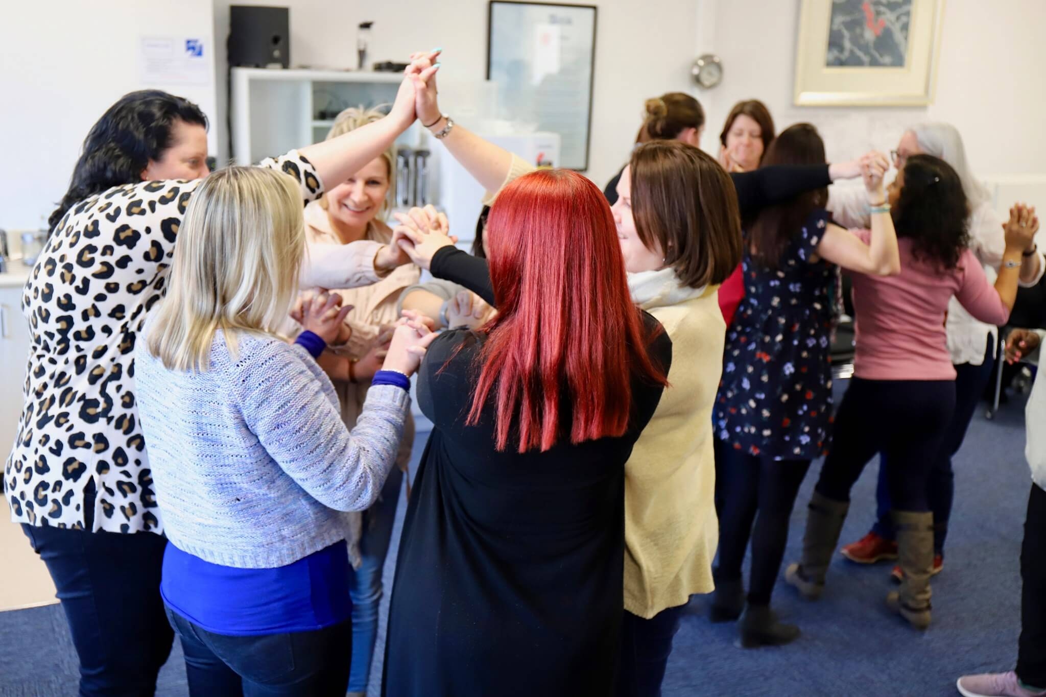 Group of diverse women participating in a team-building activity, holding hands and forming connected arches indoors.