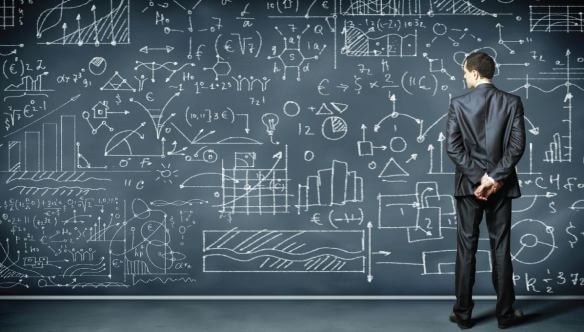 Man in a suit studies a large blackboard covered with complex mathematical equations and graphs.