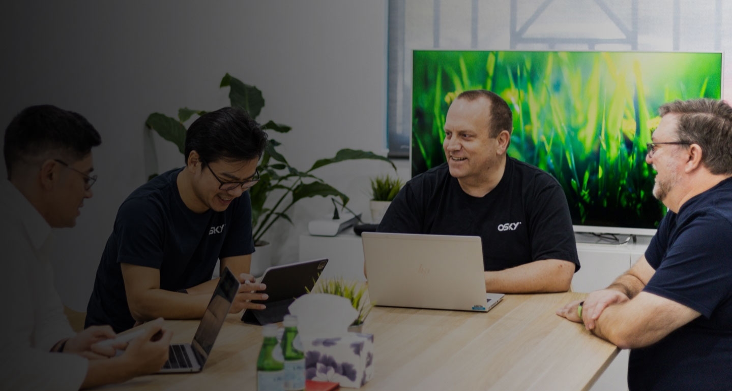 Four men smiling and working on laptops and a tablet around a conference table in an office setting.
