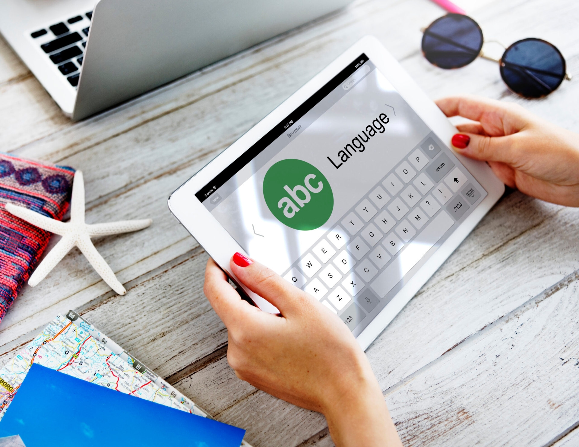 Hands using a tablet for website localization services, next to a laptop, sunglasses, and travel items on a wooden table.
