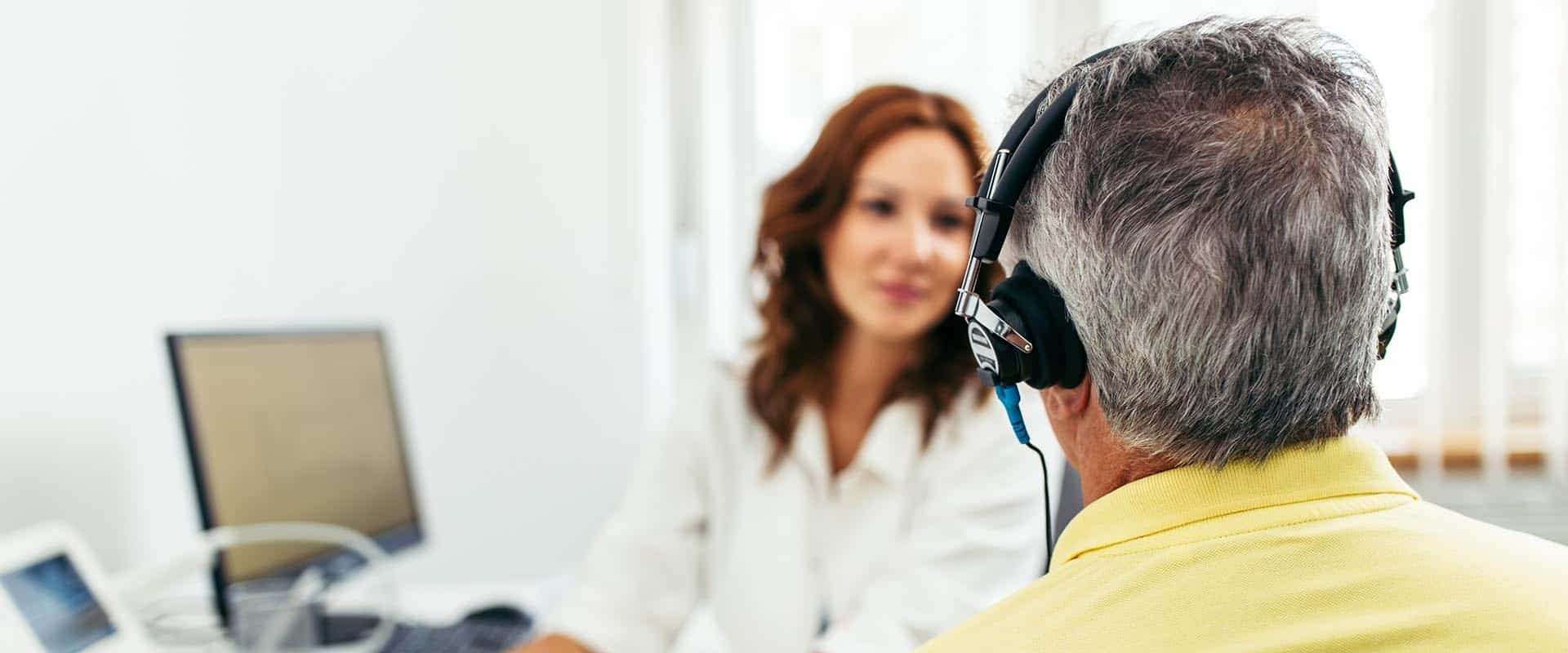 Older man wearing headphones during hearing test with female audiologist in office.